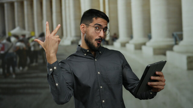 Young hispanic man gestures thumbs up while using tablet outdoors in urban setting.