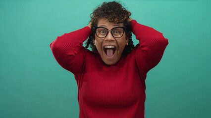 Woman with curly hair wearing red sweater and glasses holds hands on head against teal studio wall; excitement.