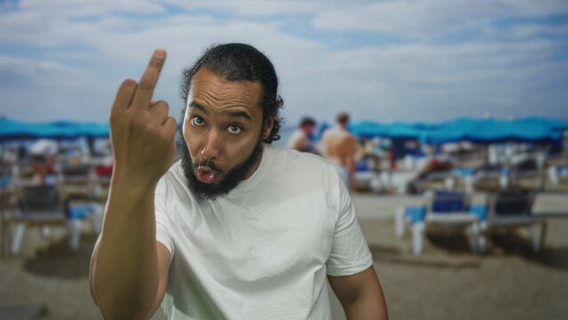 Man in white tshirt shows middle finger and grimaces while leaning forward in studio with beach chairs and blue umbrellas; defiance rebellion.