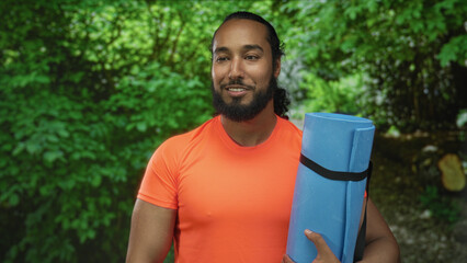 Man holding rolled blue yoga mat in forest park wearing bright orange shirt, smiling and looking...