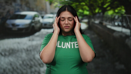 Fototapeta premium Young woman wearing green volunteer t-shirt feeling headache while standing outdoors on a city street with blurred cars and trees in the background.