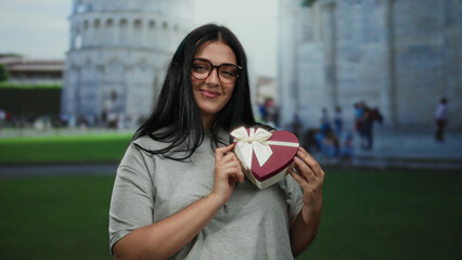 Woman smiling holding heart shaped gift box in front of pisa tower outdoors in italy showcasing...