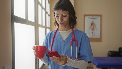Woman with stethoscope holds smartphone and red mug near window in clinic building; calm caring...