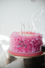 Beautiful pink ruffled birthday cake with golden candles and Happy Birthday topper against a background of balloons.