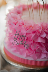 Beautiful pink ruffled birthday cake with golden candles and Happy Birthday topper against a background of balloons.