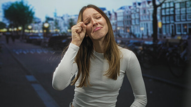 Young hispanic woman making a crying face and rubbing her eyes while leaning forward on a dim street at dusk; sadness exhaustion.