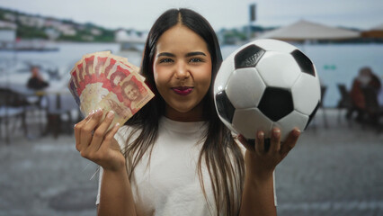 Naklejka premium Young woman holding colombian pesos and soccer ball on outdoor restaurant terrace smiling happily with a scenic backdrop.
