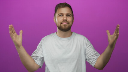 Man, bearded and smiling, shows open hands and palms in magenta studio wearing white t shirt with...