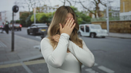 Fototapeta premium Blonde woman covers mouth with clenched fists and wide eyes on busy city street during daytime traffic rush; anxiety fear.