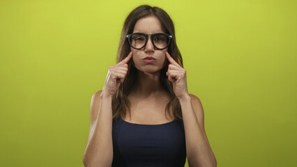 Obraz premium Young hispanic woman wearing glasses pinches puffed cheeks with pursed lips in green studio portrait; playfulness.