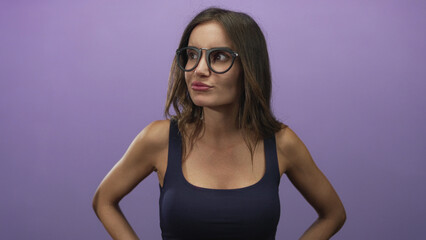 Hispanic woman wearing round glasses and black tank top with arms akimbo in purple studio; curiosity.