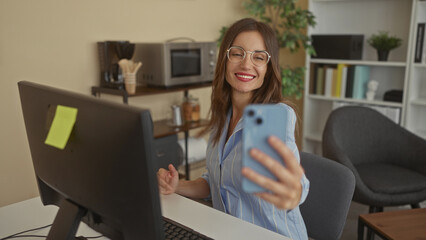 Woman wearing glasses smiles and holds smartphone for selfie at her office desk with computer monitor and bookshelves visible  confidence connection productivity. © Krakenimages.com