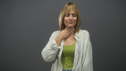 Woman touching throat and grimacing after coughing, hand at neck in studio grey backdrop wearing...