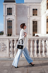 Elegant woman in white blazer and blue wide-leg trousers walking past a classical building with a stylish handbag.