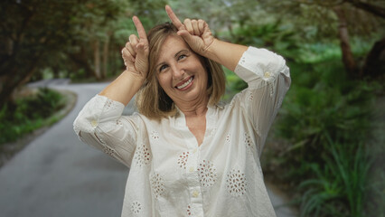 Middle-age caucasian woman holds index fingers to head while smiling broadly on street; playful joy.