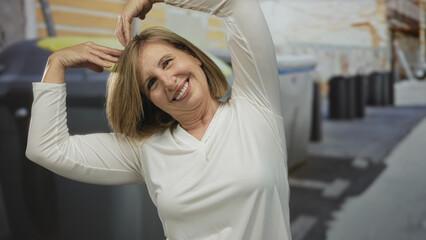 Smiling woman forms heart gesture with arms beside recycling container on city street; sustainability love.