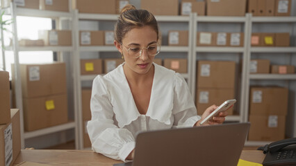 Woman holding smartphone and typing on laptop amid stacked cardboard parcels and shelving in...