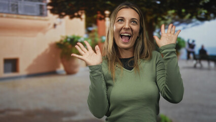 Woman smiling and waving both hands in a green sweater outdoors on a cobblestone street by a building with large planters and a bench; friendly joyful greeting.