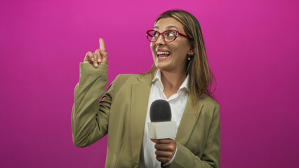 Woman reporter holding microphone points finger and laughs broadly in pink studio while wearing olive blazer; enthusiasm.