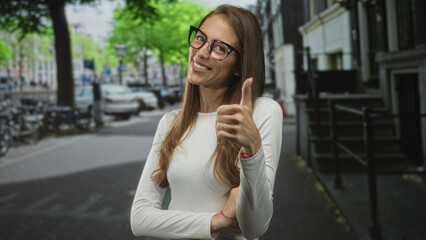 Fototapeta premium Woman wearing glasses and a white long sleeve top giving thumbs up on a busy city street, smiling with crossed arms and visible ring; confidence.