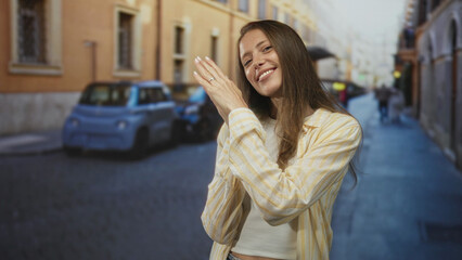 Fototapeta premium Woman clapping hands on cobbled street beside parked car with yellow building facade, smiling and showing ring; joy gratitude optimism.