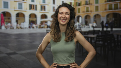 Fototapeta premium Hispanic woman in green top smiles with hands on hips in street under sunlight; happiness confidence.