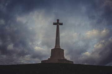 Cross of Sacrifice at British Military Cemetery in Flanders Fields. World War I CWGC memorial monument with bronze sword under dramatic cloudy sky. Western Front remembrance in Belgium.