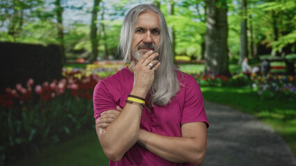 Fototapeta premium Man with hand on chin on forest path in park, long gray hair and beard, wearing magenta tshirt; contemplation thinking pondering calm.