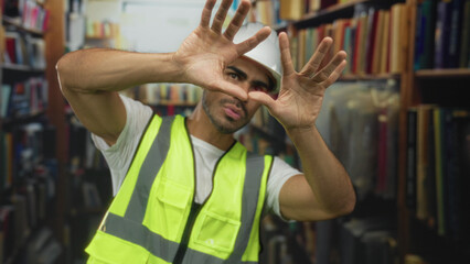Man wearing hardhat and reflective vest frames the scene with outstretched hands amid dusty bookshelves in an old library building  determination. © Krakenimages.com