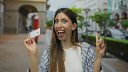 Smiling young hispanic woman holding white card with finger pointing to card on busy city street;...