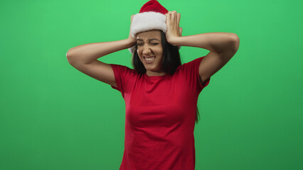Naklejka premium Young hispanic woman wearing santa hat and red shirt with hands on head and squinting face in studio on green screen; holiday frustration.