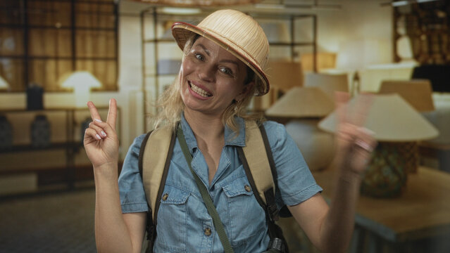 Woman wearing explorer hat and backpack makes peace sign beside lamp display in a building while smiling; joy.