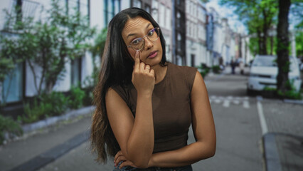 Fototapeta premium Woman touching eyeglasses with finger on cheek in street while leaning with arms crossed; pensive curiosity.