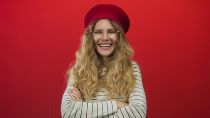 Woman with arms crossed laughing in a studio poses as a blonde young muse wearing a beret and glasses against a red striped wall.