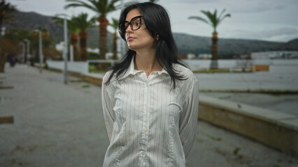 Woman wearing glasses in white shirt stands with hands behind back and turns head on street...