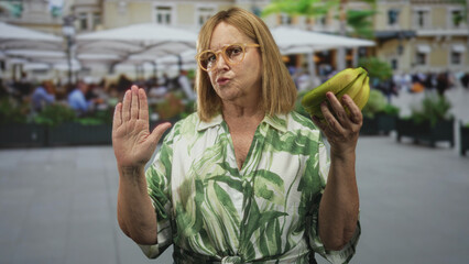 Middle age woman holds a bunch of bananas in one hand and raises an open palm in a stop gesture on a street cafe terrace with umbrellas; defiance choice resolve.