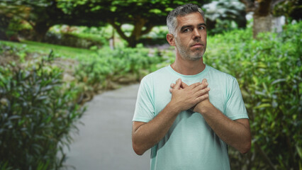Man standing with hands clasped over chest on a forest pathway wearing a mint t shirt and neutral expression; quiet contemplation.