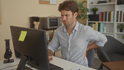 Man in striped shirt typing at desktop computer while holding lower back for backache beside office...