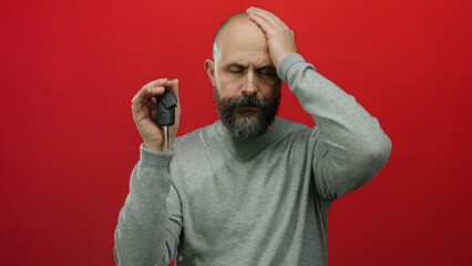 Caucasian bald man with a beard looks distressed holding car keys against a striking red...