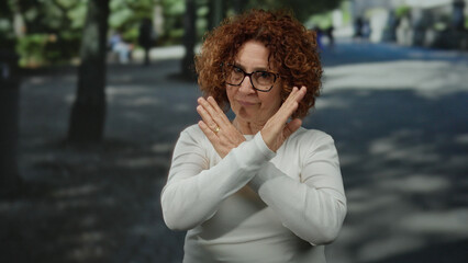 Middle-aged hispanic woman making stop gesture with fingers on a bustling city street, wearing white shirt, curly hair contrasting with outdoor urban background.