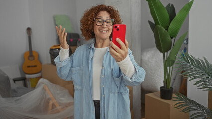 Woman smiling during video call in new home surrounded by moving boxes plants and guitar reflecting...