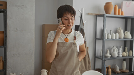 Woman wearing apron holds phone to ear among clay vessels on metal shelves in studio, short hair...