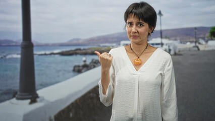 Woman pointing thumb over shoulder showing orange pendant and bare hand near chest on street by sea promenade; doubt hesitation.