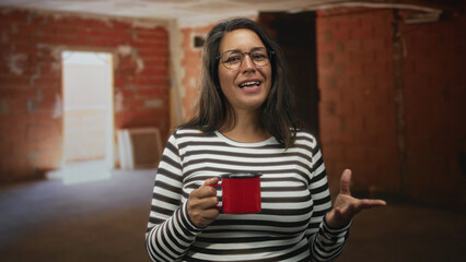 Fototapeta premium Woman holding red enamel mug with both hands and smiling, striped top visible, speaking inside an unfinished brick building interior; comfort home.