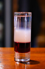 Colorful layered cocktail shot glass with orange, dark, and light layers surrounded by melting ice cubes on dark glossy bar surface, perfect for nightlife and party themes. High quality photo