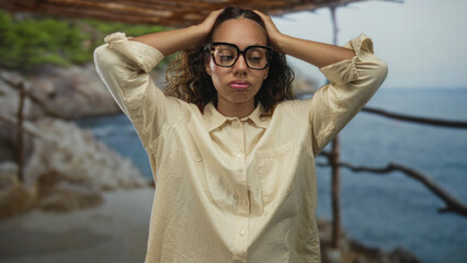 Woman with black glasses wearing beige shirt holding hands behind head on a street by the seaside under a wooden pergola; introspection.