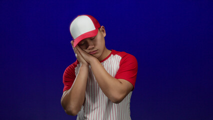 Young chinese man in baseball uniform with red and white stripes posing sleepily against isolated...