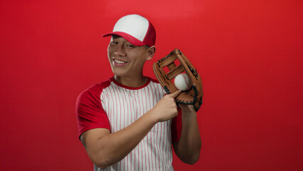 Young man in baseball uniform smiling and pointing at a baseball glove with ball against a vivid red background, showcasing sports enthusiasm and energy.