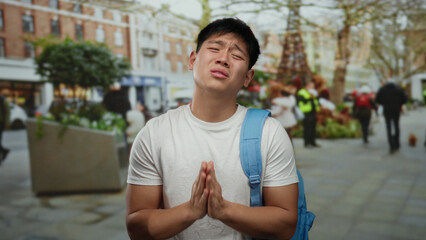 Fototapeta premium Young man with backpack standing on busy city street looking emotional with hands clasped as if praying amidst urban outdoor setting.