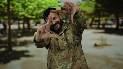 Obraz premium Hispanic man in military uniform frames his view with hands while standing on a street lined with trees, suggesting creativity outdoors.